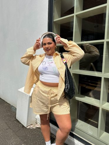 Mulher jovem de pele clara e cabelo escuto. Ela usa conjunto amarelo manteiga de camisa e bermuda, e blusa branca estampada.
