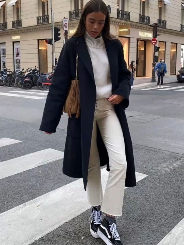 Mulher usando sobretudo azul escuro, com blusa de inverno com gola em cor branca, calça jeans em bege e vans.