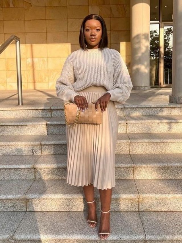 Mulher jovem e negra com cabelo curto, usando tricô, saia plissada e sandália de salto.