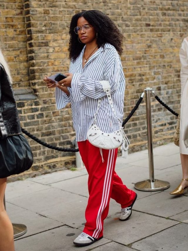 Mulher negra, usando camisa azul listrada oversized, segurando uma bolsa de ombro em cor branca e usando calça Adidas vermelha.