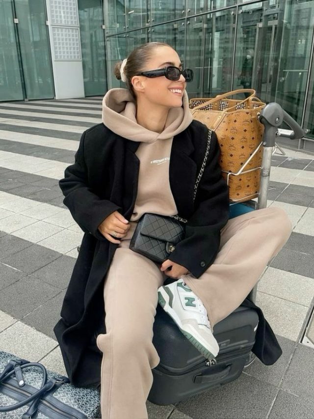 Mulher loira e jovem, sentada em mala de viagem, usando o cabelo em coque e óculos de sol, usando moletom de capuz em cor bege, calça de moletom na mesma cor, sobretudo preto, bolsa pequena em cor preta cruzada ao corpo e tênis branco.