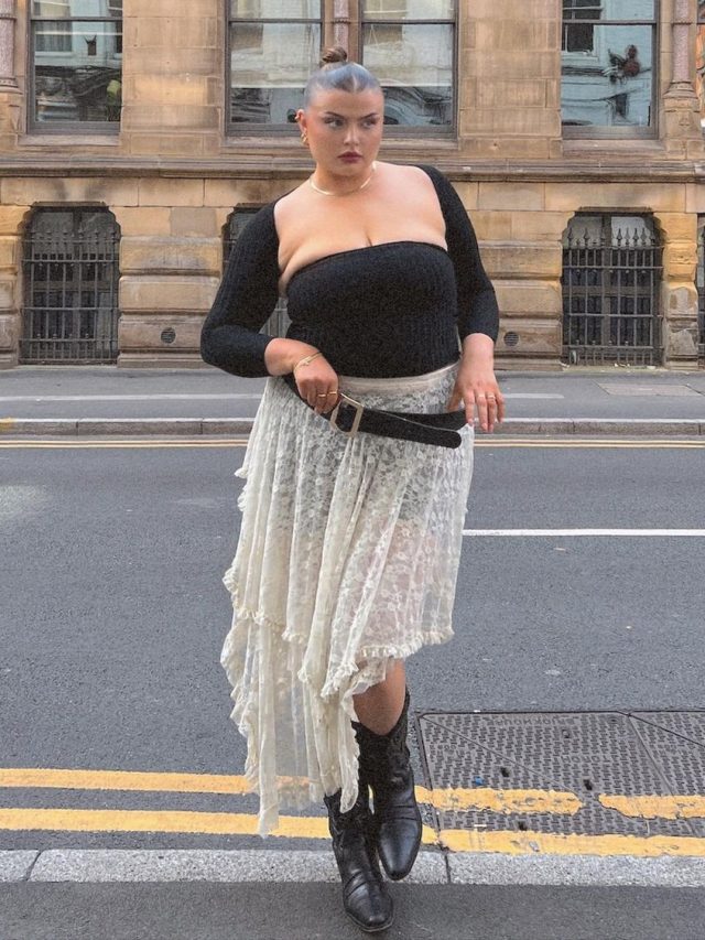 Francesca Perks com blusa preta, saia de renda branca e bota preta