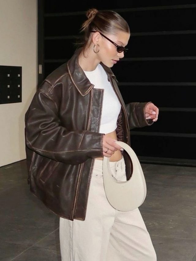 Mulher jovem de pele clara. Ela veste roupa bege e jaqueta de couro marrom.