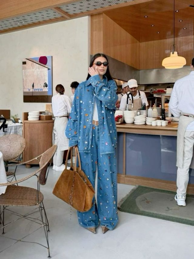 Mulher jovem de pele clara com cabelo escuro. Ela usa óculos e look all jeans com pedrarias.