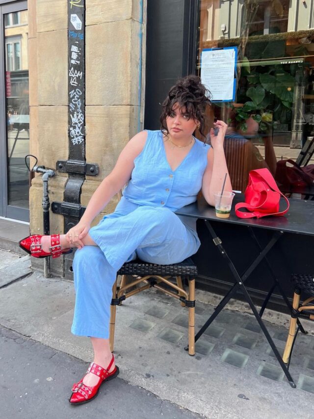 Mulher jovem e midsize com pele clara. Ela usa conjunto azul claro e acessórios vermelhos (sapatilha e bolsa).