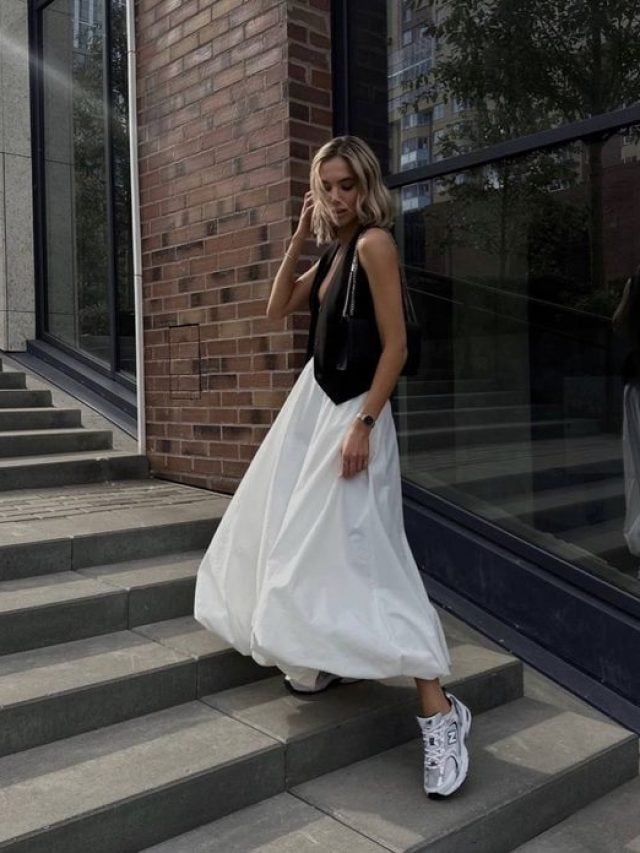 Mulher jovem e magra com pele clara. Ela veste colete de alfaiataria preto e saia balonê branca com tênis branco