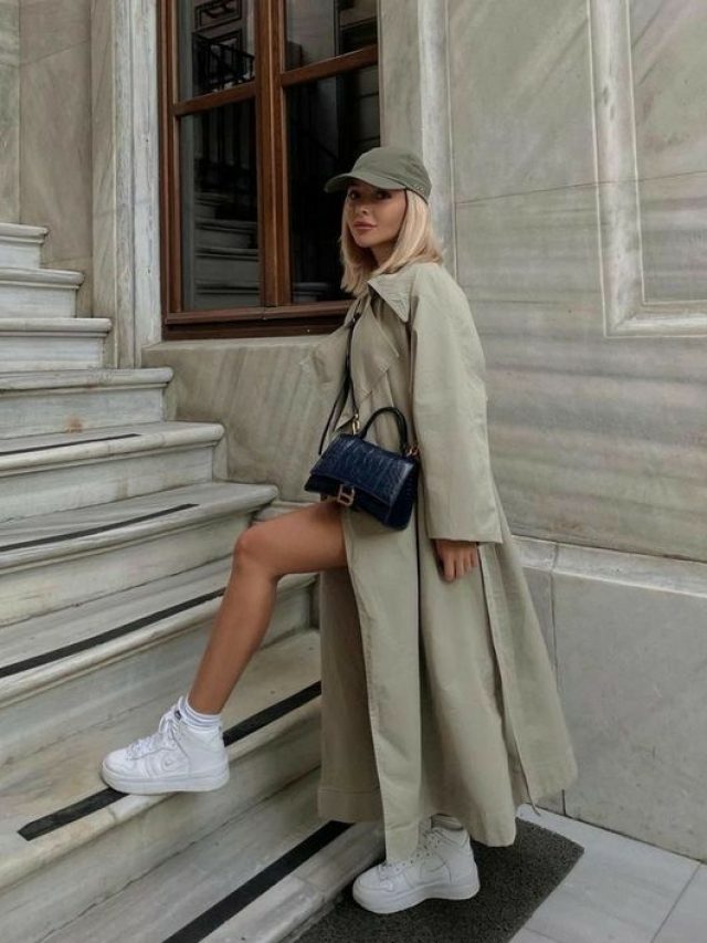 Mulher jovem de pele clara usando boné verde, trench coat e tênis branco.