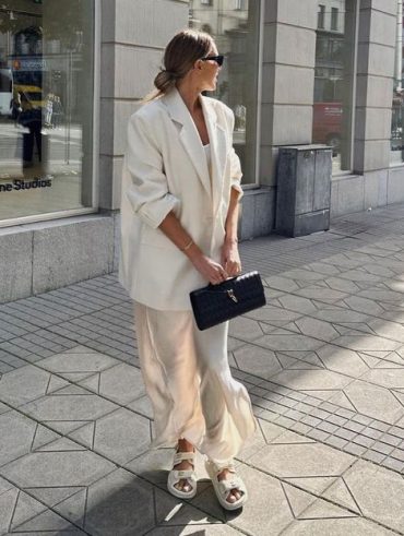 Mulher com coque, usando óculos de sol, blazer branco e vestido de cetim.
