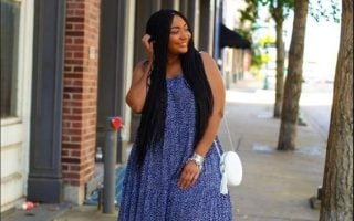 Mulher jovem e negra com cabelo longo, usando vestido longo em tonalidade azul, bolsa branca no ombro e tênis.