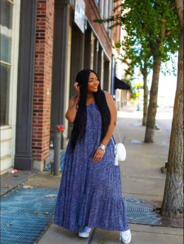 Mulher jovem e negra com cabelo longo, usando vestido longo em tonalidade azul, bolsa branca no ombro e tênis.