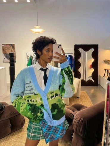Mulher negra e jovem. Ela está em frente ao espelho em foto. Ela veste cardigan estampado nas cores azul e verde, camisa branca e gravata preta.