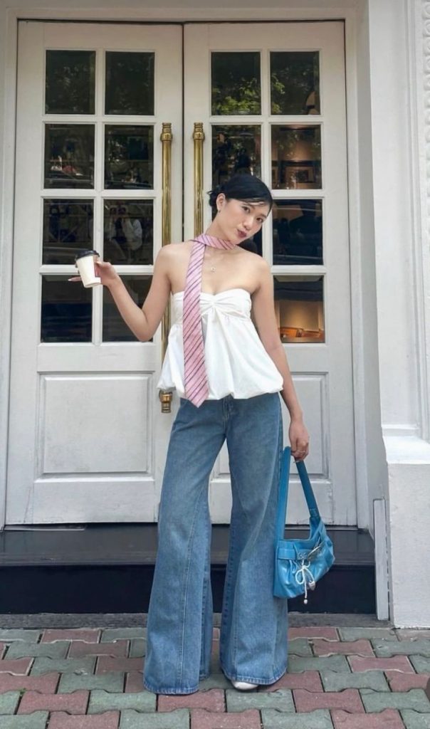 Mulher jovem de pele clara. Ela veste look com acessórios, calça jeans e blusa branca. 