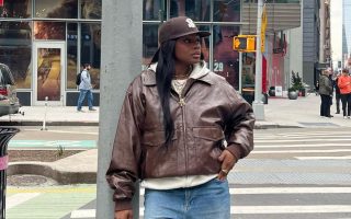 Mulher jovem de pele negra. Ela veste jaqueta de couro marrom e calça jeans clara.