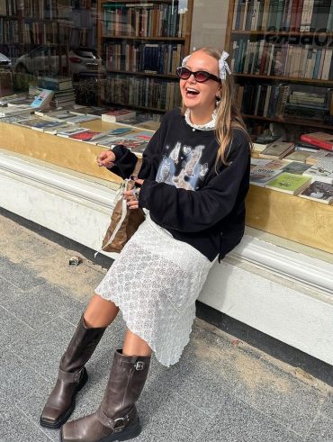 Mulher jovem e magra de pele clara com cabelo loiro liso. Ela usa botas na cor marrom, saia de renda e moletom.