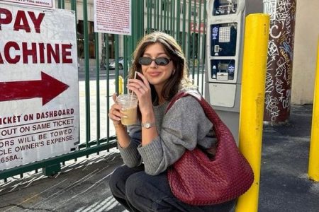 Mulher jovem de pele clara e cabelos castanhos. Ela usa suéter cinza, bolsa de ombro grande vermelha, calça jeans escura e sapatilha vermelha.