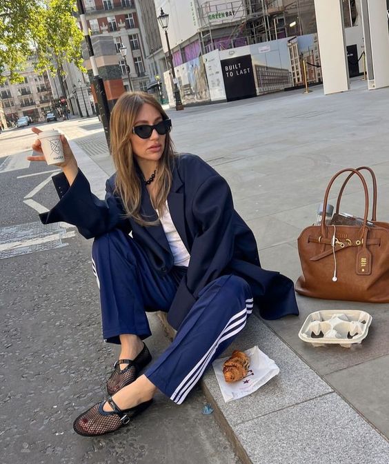 Mulher branca usando blazer preto, calça Adidas azul e sapatilha telada