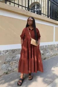 Mulher negra e com cabelo longo com tranças, usando óculos de sol, vestido marrom no modelo três marias, papete e uma bolsa bege.