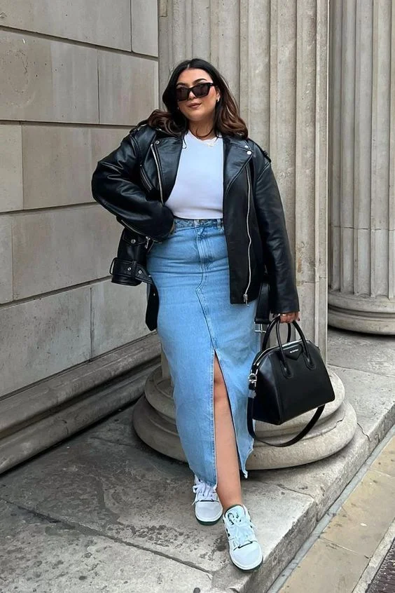 Mulher jovem, branca, plus size com cabelo moreno, usando óculos de sol, jaqueta de couro, blusa branca, saia jeans com fenda e tênis branco.