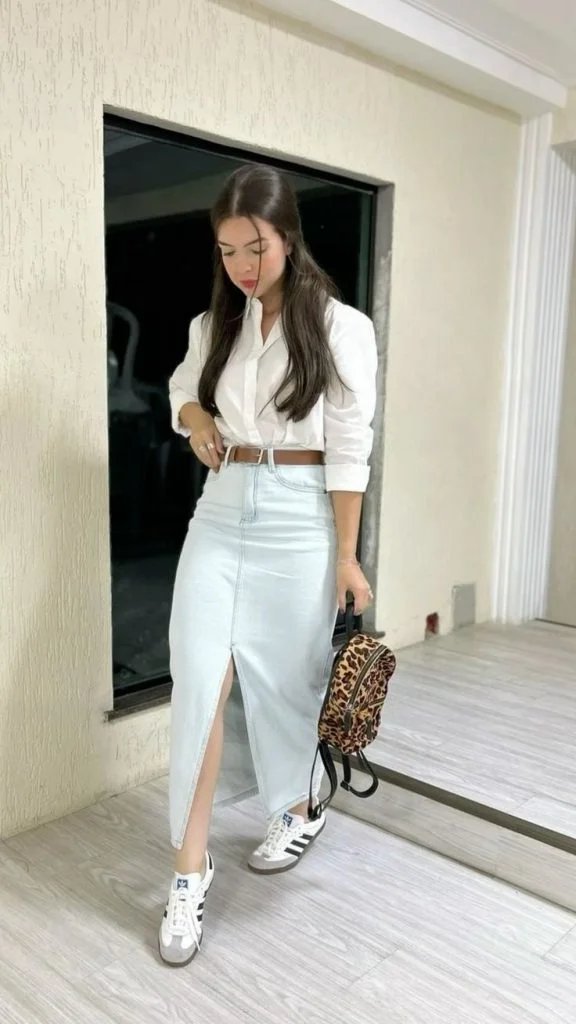 Mulher jovem e branca, com cabelo longo e moreno, usando camisa branca, saia jeans longa com fenda, cinto marrom, calçando Adidas Samba e segurando uma bolsa de oncinha.