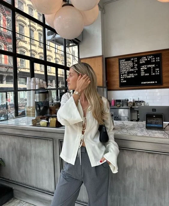 Mulher jovem e com cabelo loiro, usando blusa boho com abertura de amarrações frontais, calça de alfaiataria na cor cinza e bolsa pequena em cor preta no ombro.