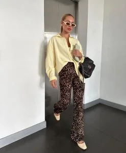 Mulher loira e branca, com cabelo preso, usando óculos de sol, uma camisa em amarelo-manteiga, calça de oncinha e sapatilha.