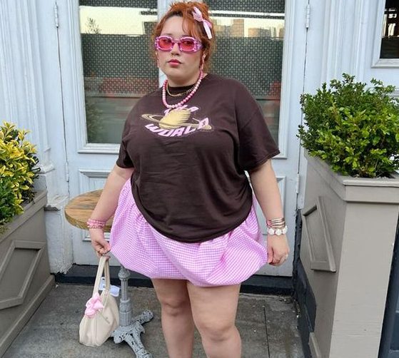 Mulher plus size e branca, com cabelo preso em cor ruivo e com acessório de laço, usando óculos de sol rosa, camiseta oversized em cor marrom, saia balonê curta em rosa, segurando uma bolsa pequena em cor rosa e usando tênis branco com detalhes rosa.