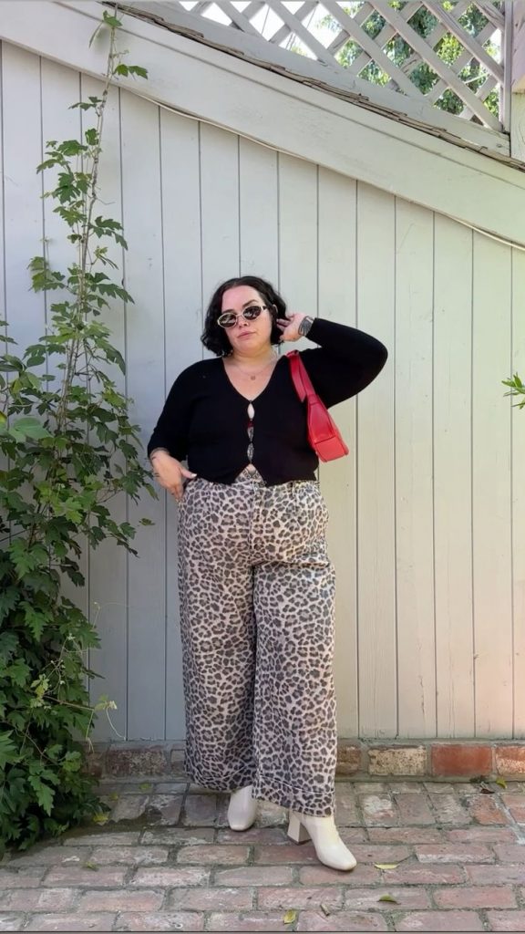 Mulher jovem, branca e plus size, com cabelo curto, usando um cardigan preto, bolsa de ombro em cor vermelha, calça de oncinha e bota de cano baixo de salto em cor branca.