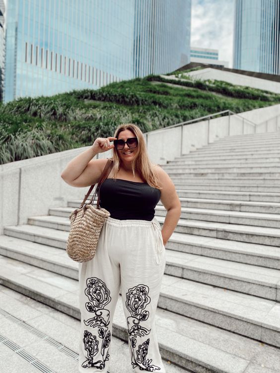 Mulher jovem, branca e gorda, com cabelo loiro, usando óculos de sol, blusa sem alça em cor preta e calça branca com estampa preta.