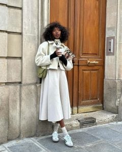 Mulher negra e jovem, segurando copo de café, usando look neutro com peças em branco e calçando Adidas Samba.