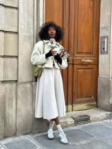 Mulher negra e jovem, segurando copo de café, usando look neutro com peças em branco e calçando Adidas Samba.
