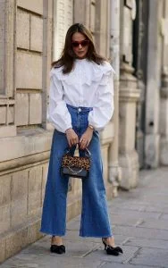 Mulher jovem e com cabelo médio em castanho claro, usando óculos de sol, camisa branca, calça jeans e segurando uma bolsa.