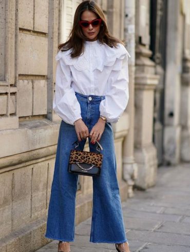 Mulher jovem e com cabelo médio em castanho claro, usando óculos de sol, camisa branca, calça jeans e segurando uma bolsa.
