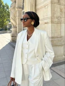 Mulher jovem, negra e magra, usando cabelo preso, óculos de sol, look monocromático branco com colete, blazer e calça de alfaiataria.