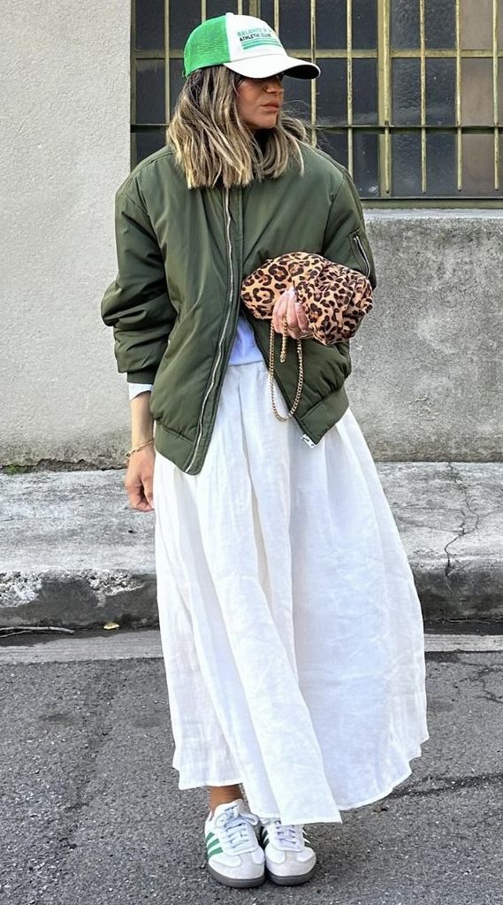 Mulher jovem e loira com cabelo curto, usando boné em cor verde, jaqueta verde, saia longa branca e Adidas Samba.