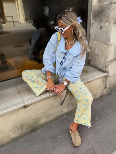 Mulher branca usando óculos de sol branco, piranha de flor no cabelo, colar de corrente, brinco de corrente, blusa azul-claro com amarração frontal, calça estampada e mule Birkenstock