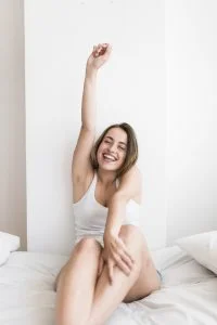 Mulher branca com cabelo curto e castanho, usando regata branca e short, e sorrindo com um braço levantado e a axila à mostra