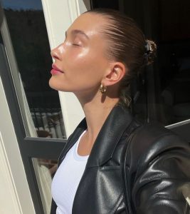 Hailey Bieber com blusa branca, jaqueta de couro, cabelo em coque e pele perfeita e brilhante