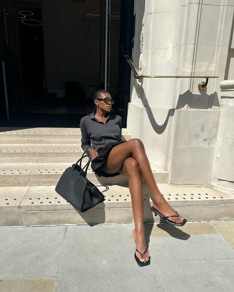 Georgia usando óculos de sol, maxi bag preta, blusa gola polo cinza, saia balonê preta e chinelo kitten heels preto