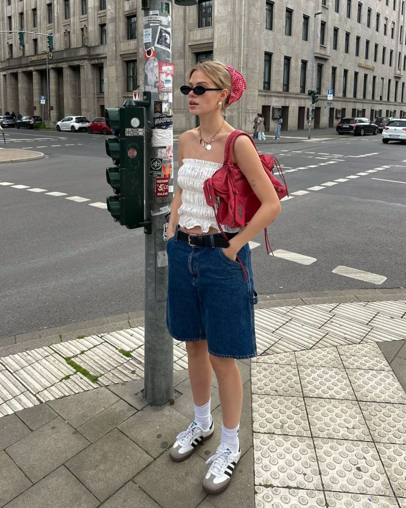 Linda com acessório de cabelo, óculos de sol, blusa branca sem alça, bolsa vermelha, jorts, meia branca e Adidas Samba