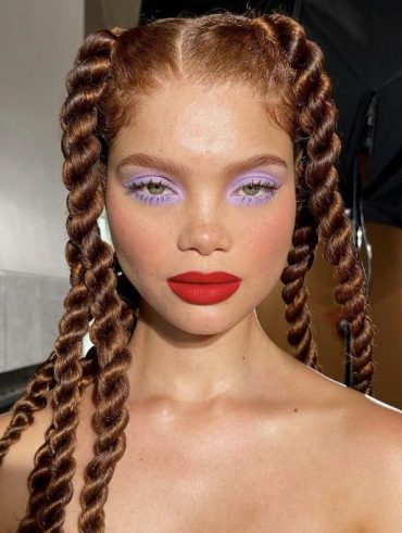 Mulher com cabelo ruivo trançado, batom vermelho e sombra lilás