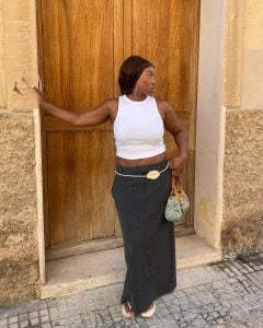 Nnenna usando regata branca, saia midi risca de giz cinza, cinto de flor e chinelo de dedo