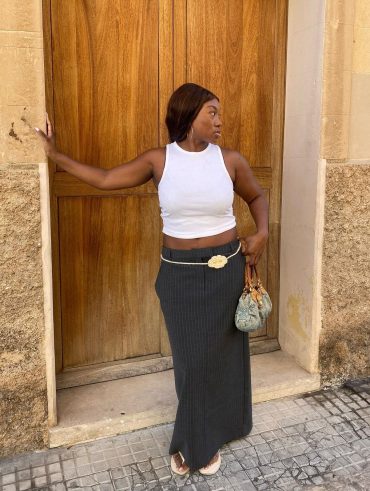 Nnenna usando regata branca, saia midi risca de giz cinza, cinto de flor e chinelo de dedo