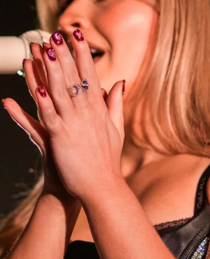 Sabrina Carpenter com a mão encostada, com anéis e unha curtinha vermelha com corações vazados e strass