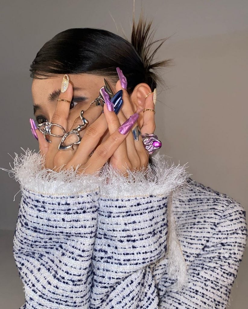 Mulher branca com cabelo preso, anéis prata e unhas maximalistas
