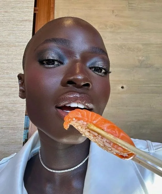 Mulher negra com blusa branca, comendo sushi e com maquiagem metálica