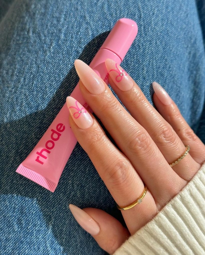 Mão branca com anéis dourados e unha amendoada brilhosa com laço rosa
