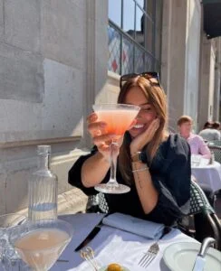 Mulher jovem com taça com drink. Ela usa óculos de ol na cabeça e blusa preta.