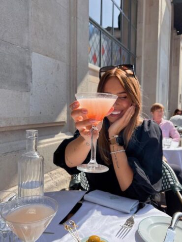 Mulher jovem com taça com drink. Ela usa óculos de ol na cabeça e blusa preta.