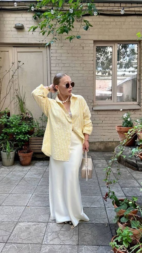 Mulher jovem e magra com pele clara. Ela usa camisa amarelo manteiga, saia de cetim e colar de pérolas.