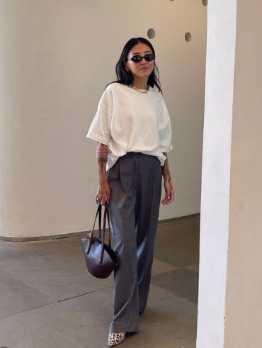 Mulher jovem de pele clara e cabelo escuro. Ela usa óculos de sol e bolsa. Ela veste camiseta branca e calça cinza.